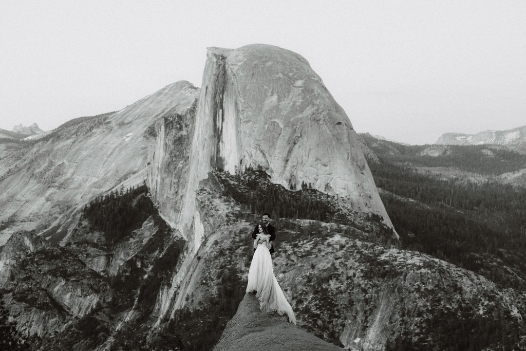 A bride and groom stand on a rocky ledge overlooking Half Dome in Yosemite National Park, surrounded by mountains and forest for a last minute elopement in Yosemite
