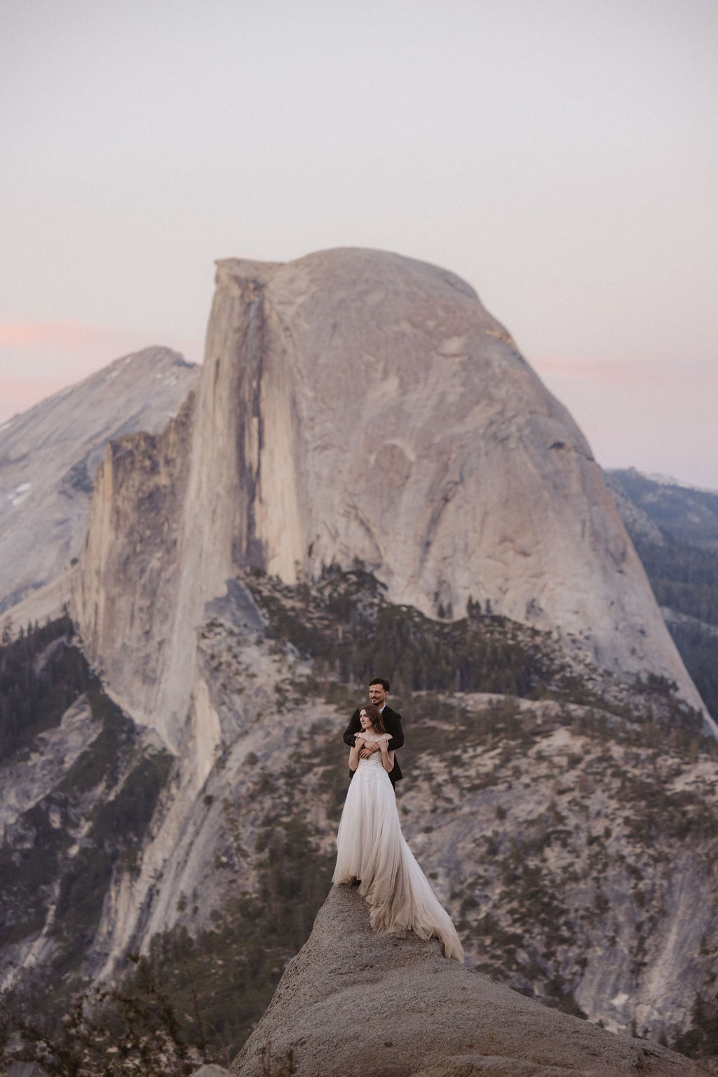 A bride and groom stand on a rocky ledge overlooking Half Dome in Yosemite National Park, surrounded by mountains and forest for a last minute elopement in Yosemite