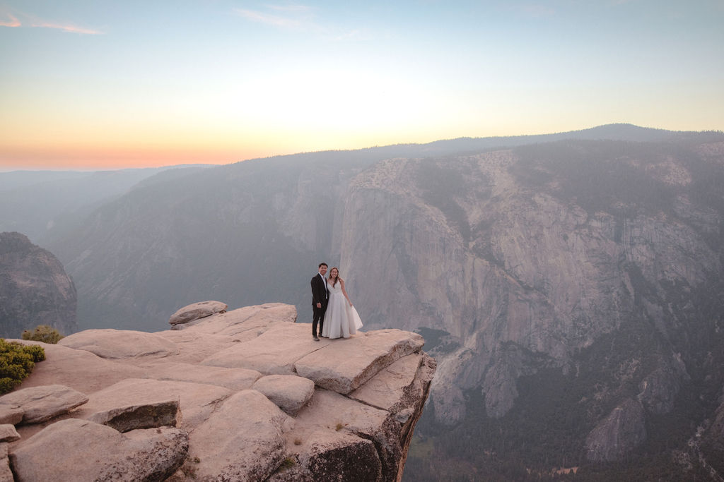 A bride and groom stand together on a rocky cliff edge at sunset with mountains and a wide valley in the background.