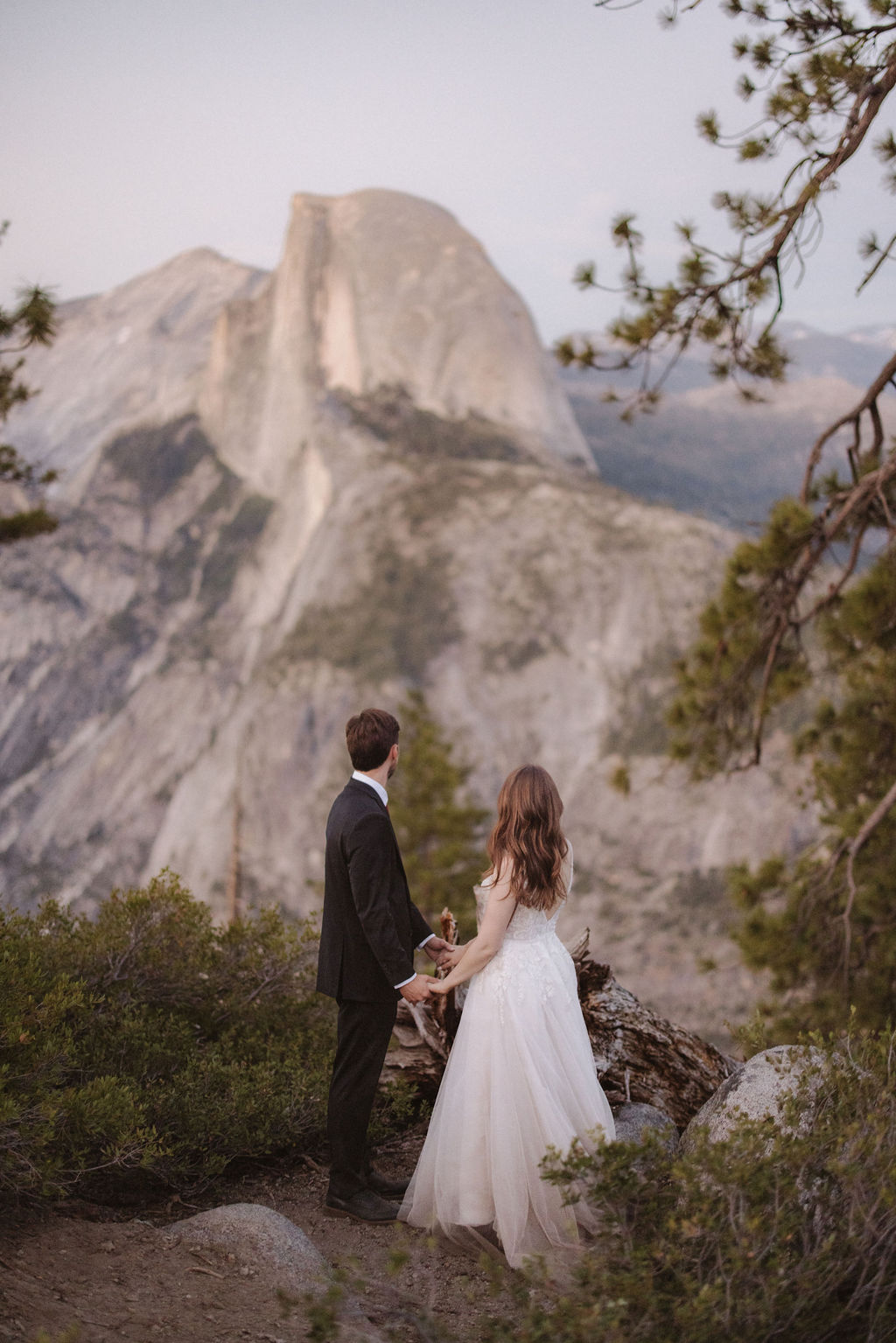 A bride and groom stand on a rocky ledge overlooking Half Dome in Yosemite National Park, surrounded by mountains and forest for a last minute elopement in Yosemite