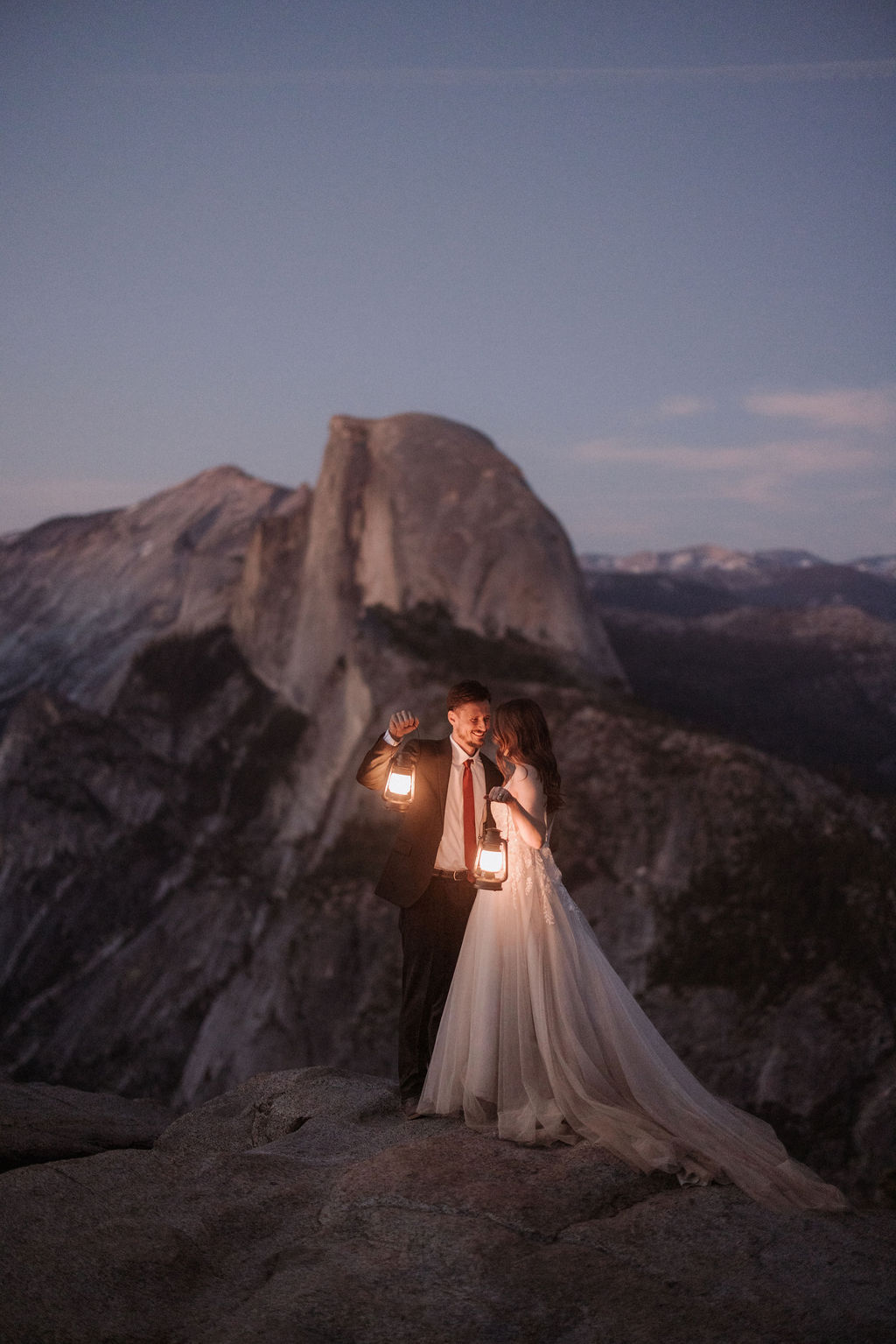 A bride and groom stand on a rocky outcrop holding lanterns, with a large mountain peak and a twilight sky in the background.