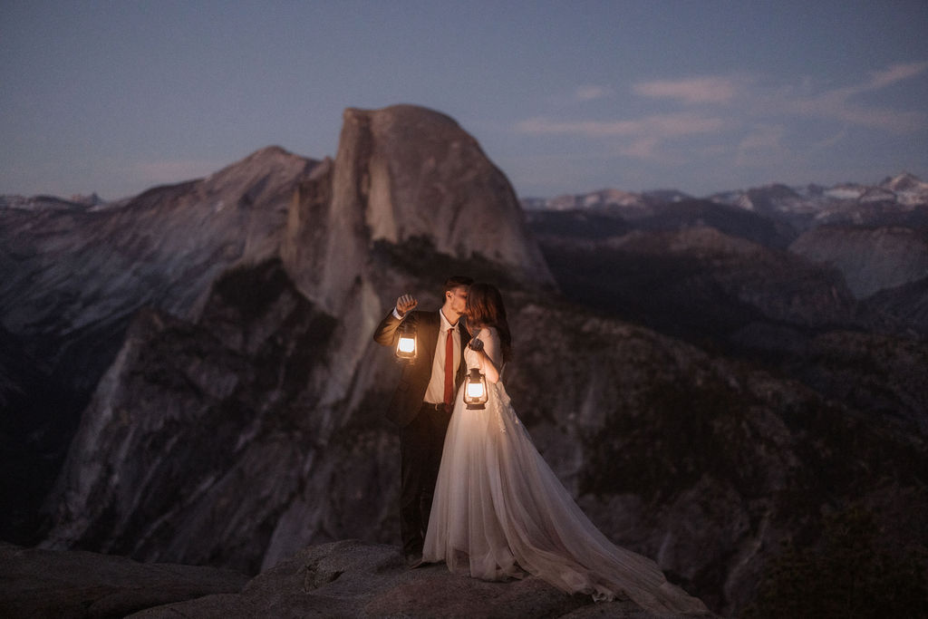 A bride and groom stand on a rocky ledge holding lanterns, with a mountain and twilight sky in the background for a last minute elopement in Yosemite