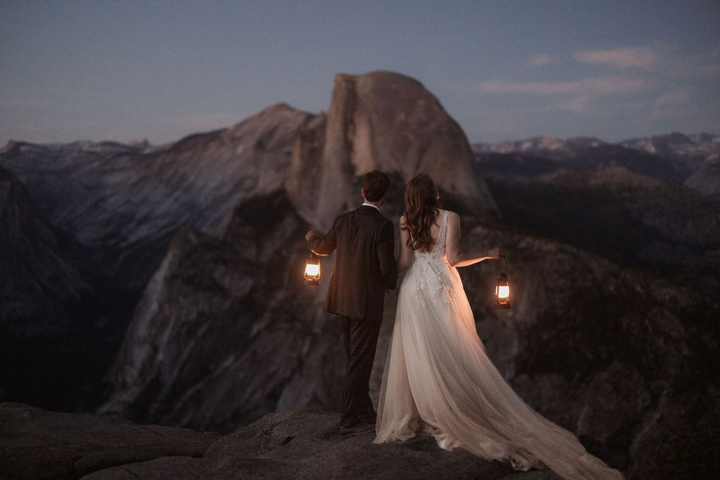 A bride and groom stand on a rocky ledge holding lanterns, with a mountain and twilight sky in the background for a last minute elopement in Yosemite
