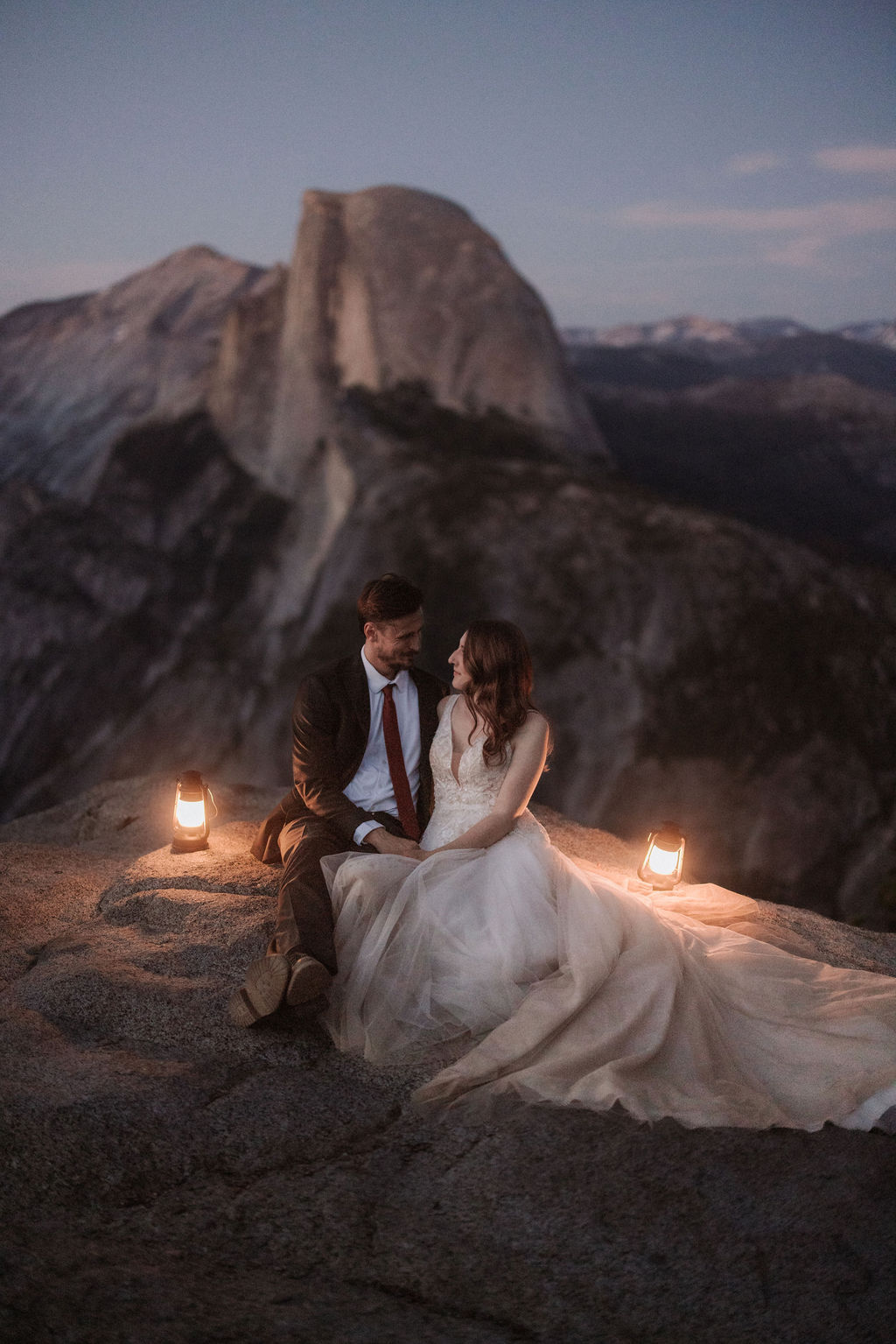 A bride and groom stand on a rocky outcrop holding lanterns, with a large mountain peak and a twilight sky in the background for their last minute elopement in Yosemite