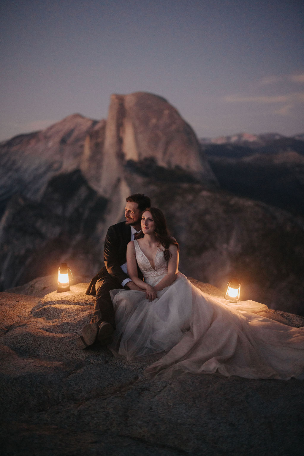 A bride and groom stand on a rocky ledge holding lanterns, with a mountain and twilight sky in the background for a last minute elopement in Yosemite