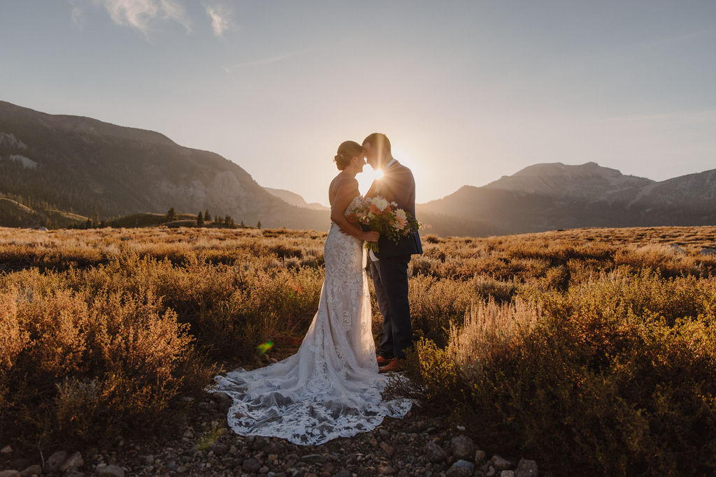 A bride and groom stand together in a sunlit field with mountains in the background during sunset.
