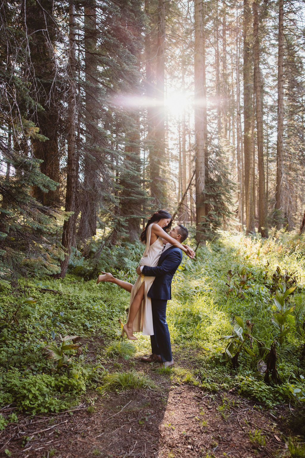 A couple embraces in a sunlit forest, with the woman lifting one leg as she is hugged by the man amid tall trees and green foliage for their post elopement session