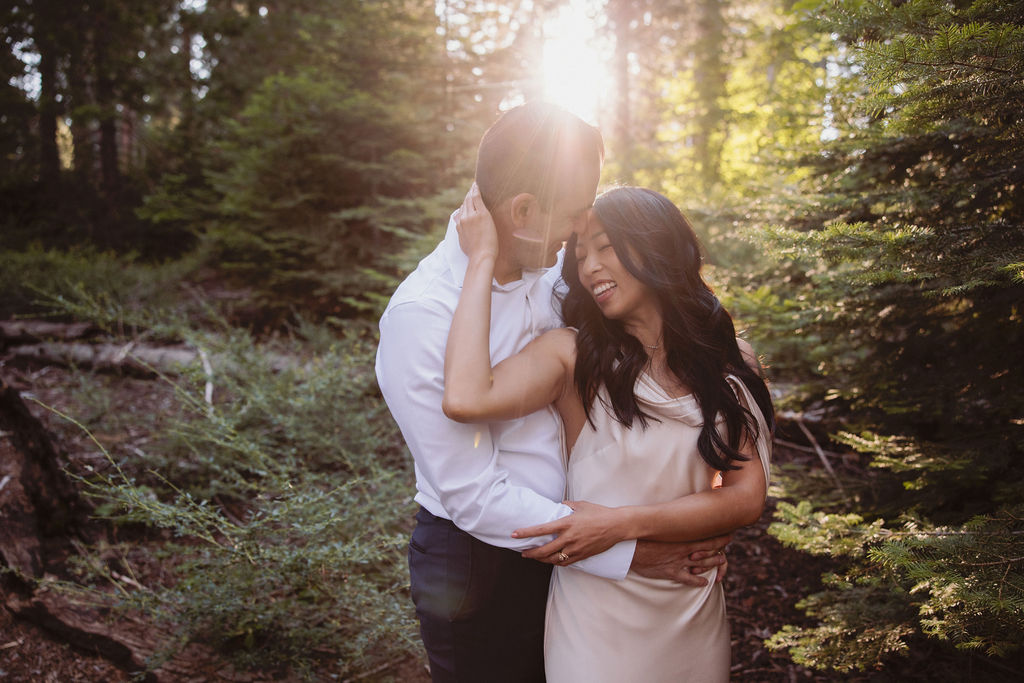 A couple embraces in a sunlit forest, with the woman lifting one leg as she is hugged by the man amid tall trees and green foliage for their post elopement session