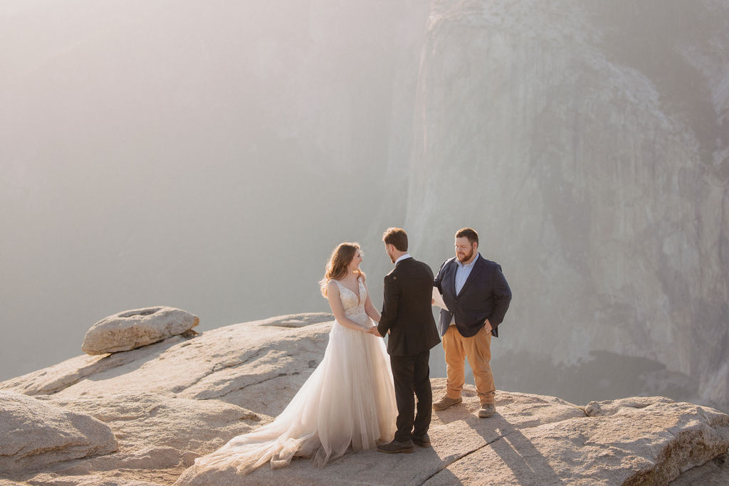 A bride, groom, and officiant stand on the edge of a rocky cliff at sunrise or sunset with mountains in the background for a last minute elopement in Yosemite