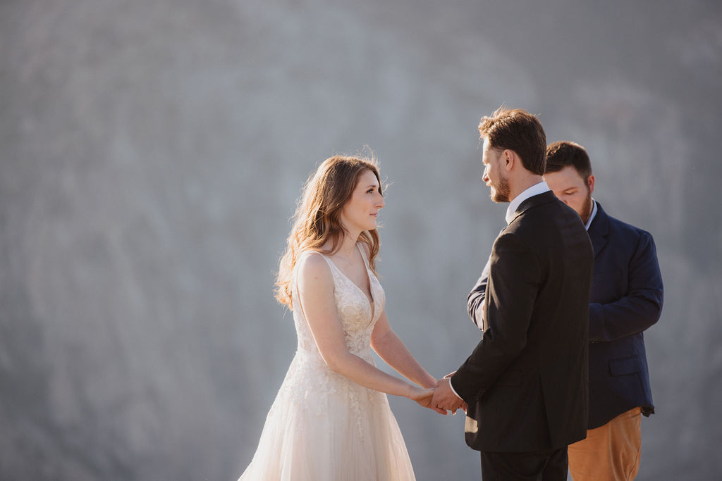 A bride, groom, and officiant stand on the edge of a rocky cliff at sunrise or sunset with mountains in the background for a last minute elopement in Yosemite