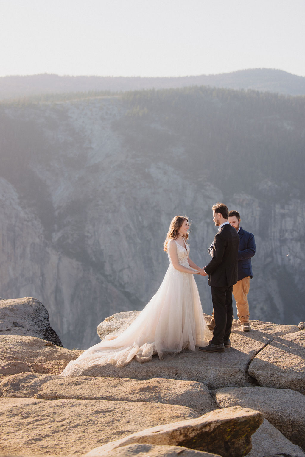 A bride, groom, and officiant stand on the edge of a rocky cliff at sunrise or sunset with mountains in the background for a last minute elopement in Yosemite