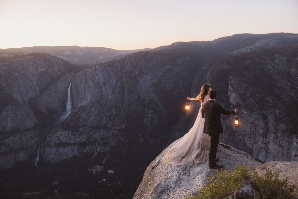 A bride and groom stand on a rocky ledge holding lanterns, with a mountain and twilight sky in the background for a last minute elopement in Yosemite