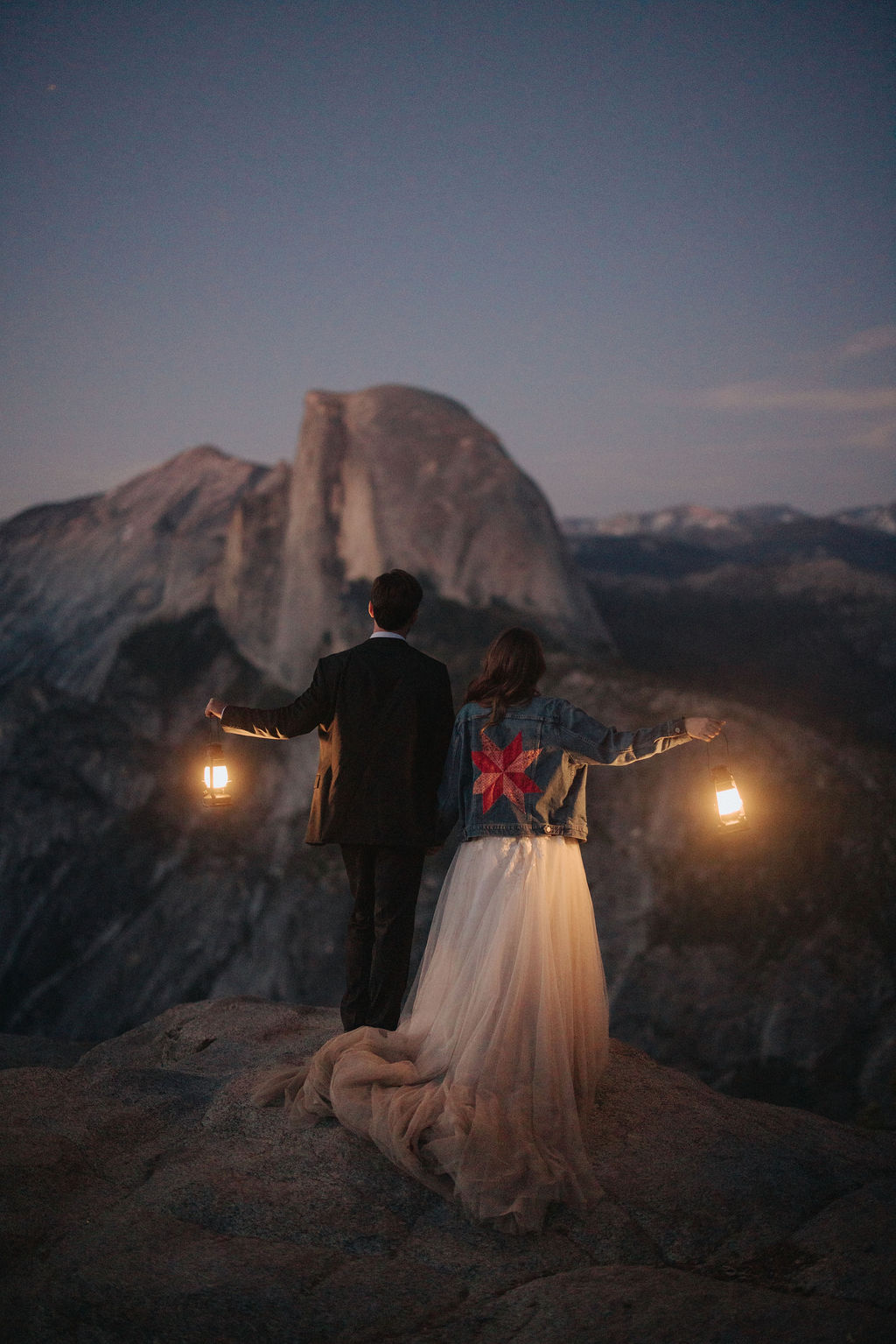 A bride and groom stand on a rocky ledge holding lanterns, with a mountain and twilight sky in the background for a last minute elopement in Yosemite