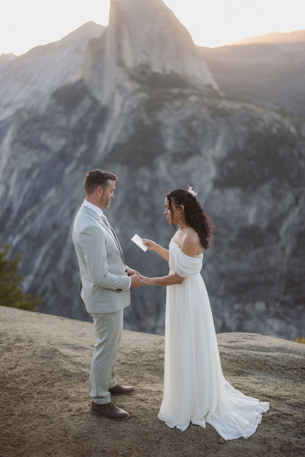 A bride and groom kiss outdoors on a rocky ledge with a mountainous landscape in the background.