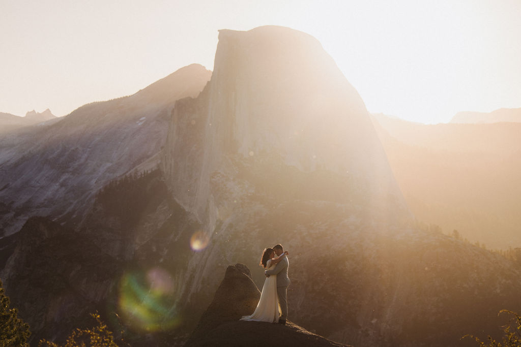 A couple embraces on a rocky ledge with a large mountain and bright sunlight in the background.