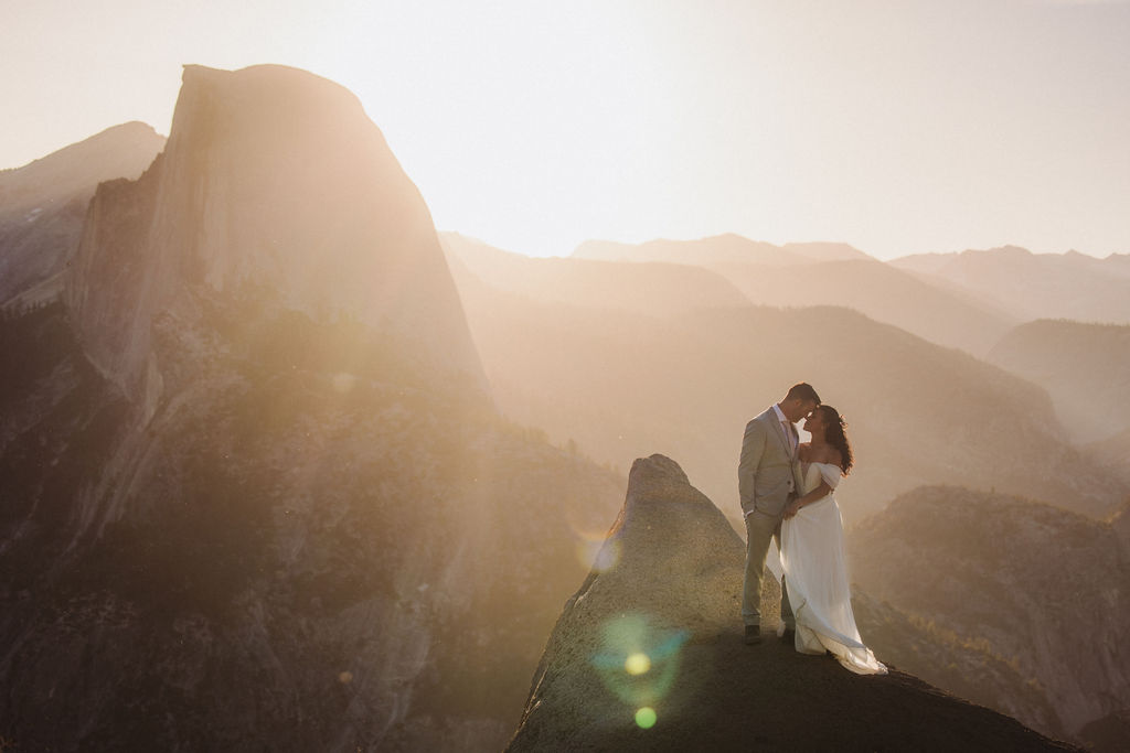 A bride and groom stand on a rocky mountaintop at sunset, sharing an intimate moment with mountains and sunlight in the background for their elopement adventure
