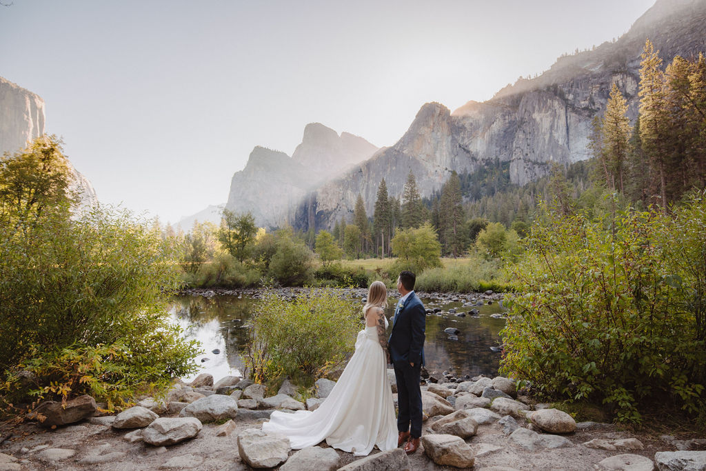 A bride and groom stand on rocky ground by a calm river, surrounded by trees, with mountains in the background under a clear sky.
