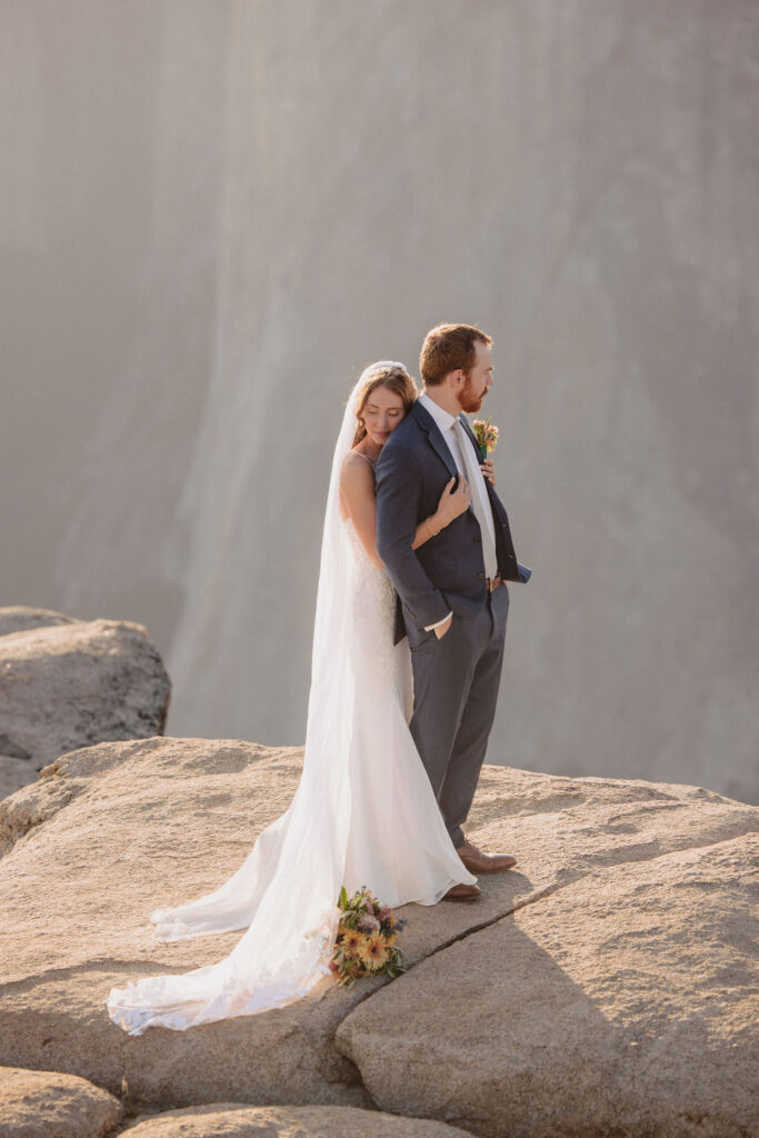 A bride and groom kiss on a rocky cliff with a mountainous background, dressed in formal wedding attire, under soft natural light by an experienced elopement photographer