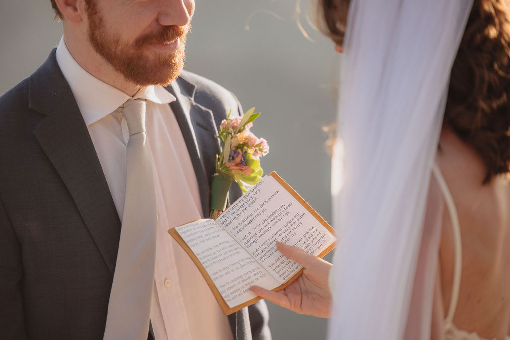 A bride and groom kiss on a rocky cliff with a mountainous background, dressed in formal wedding attire, under soft natural light by an experienced elopement photographer