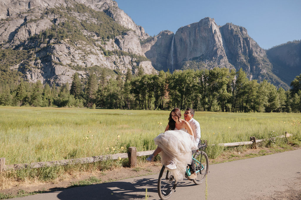 Bride and groom ride a tandem bicycle on a paved path with mountains and green meadows in the background under a clear blue sky.