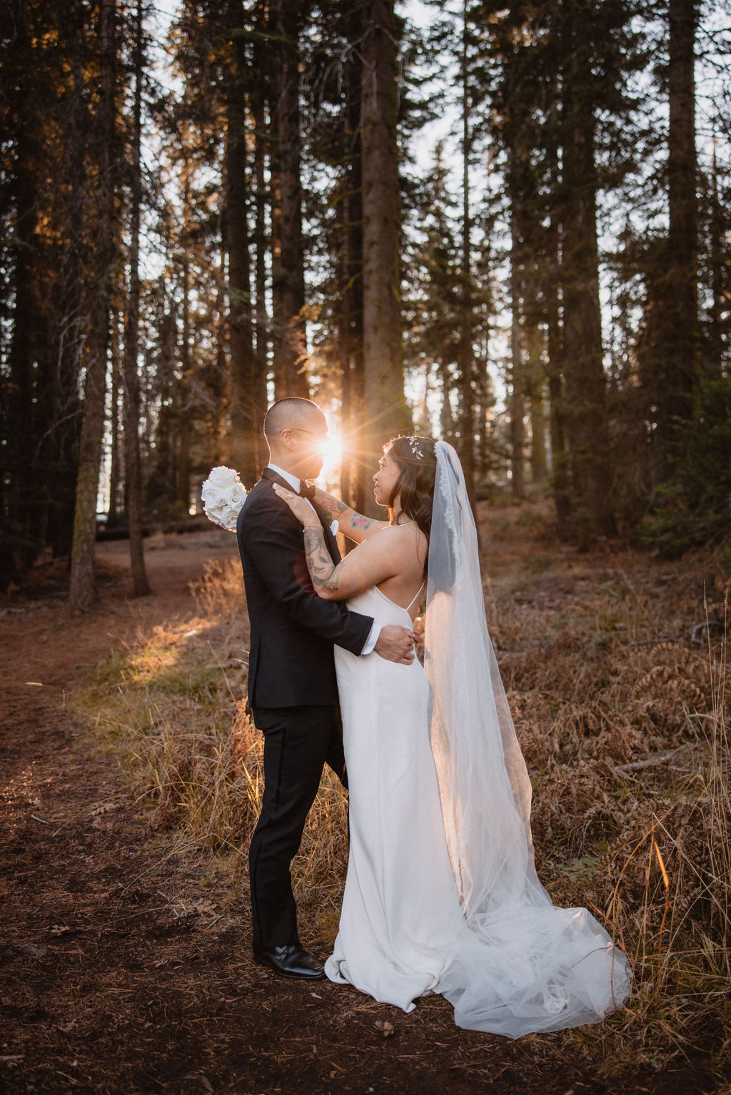 A bride and groom stand together at the base of giant sequoia trees in a forest, holding hands and looking up at the trees for an adventure elopement
