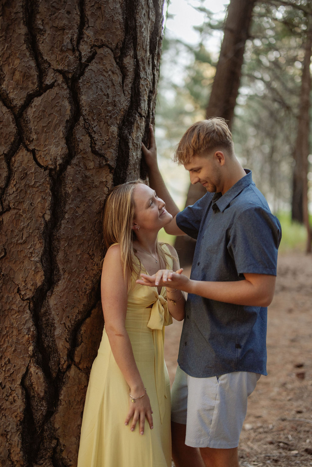 A woman in a yellow dress and a man in a blue shirt stand by a tree in a forest, looking at each other and smiling.