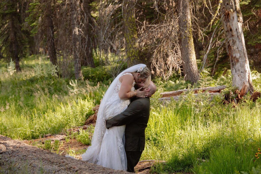 A bride in a white dress and veil is lifted and kissed by a groom in a suit in a forested, grassy area.