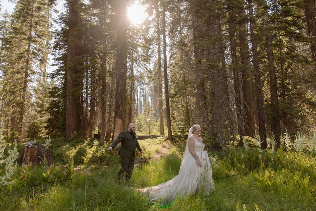 A bride in a white dress and veil is lifted and kissed by a groom in a suit in a forested, grassy area.