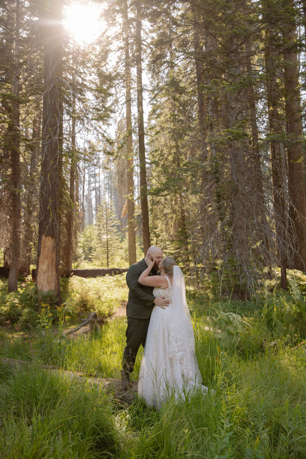 A bride in a white dress and veil is lifted and kissed by a groom in a suit in a forested, grassy area.