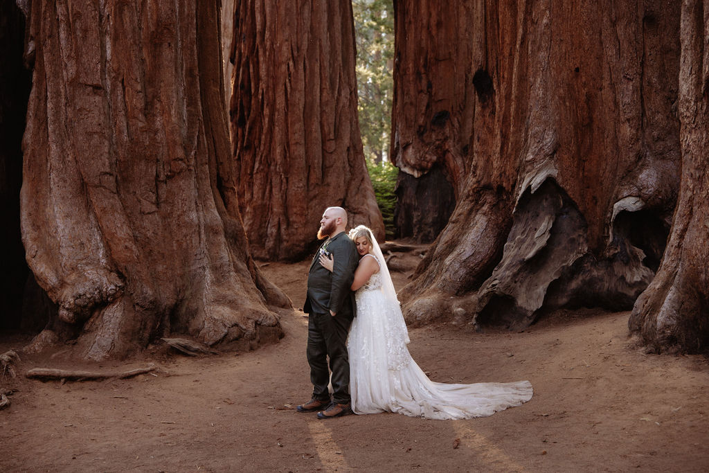 A bride and groom stand holding hands in front of giant redwood trees in a forest for their elopement in Yosemite and Sequoia