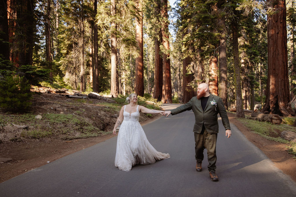 A bride in a white dress and a groom in a green suit walk hand in hand on a paved road through a forest of tall trees.