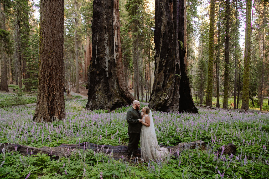 A bride and groom stand together in a forest clearing surrounded by tall trees and purple wildflowers.
