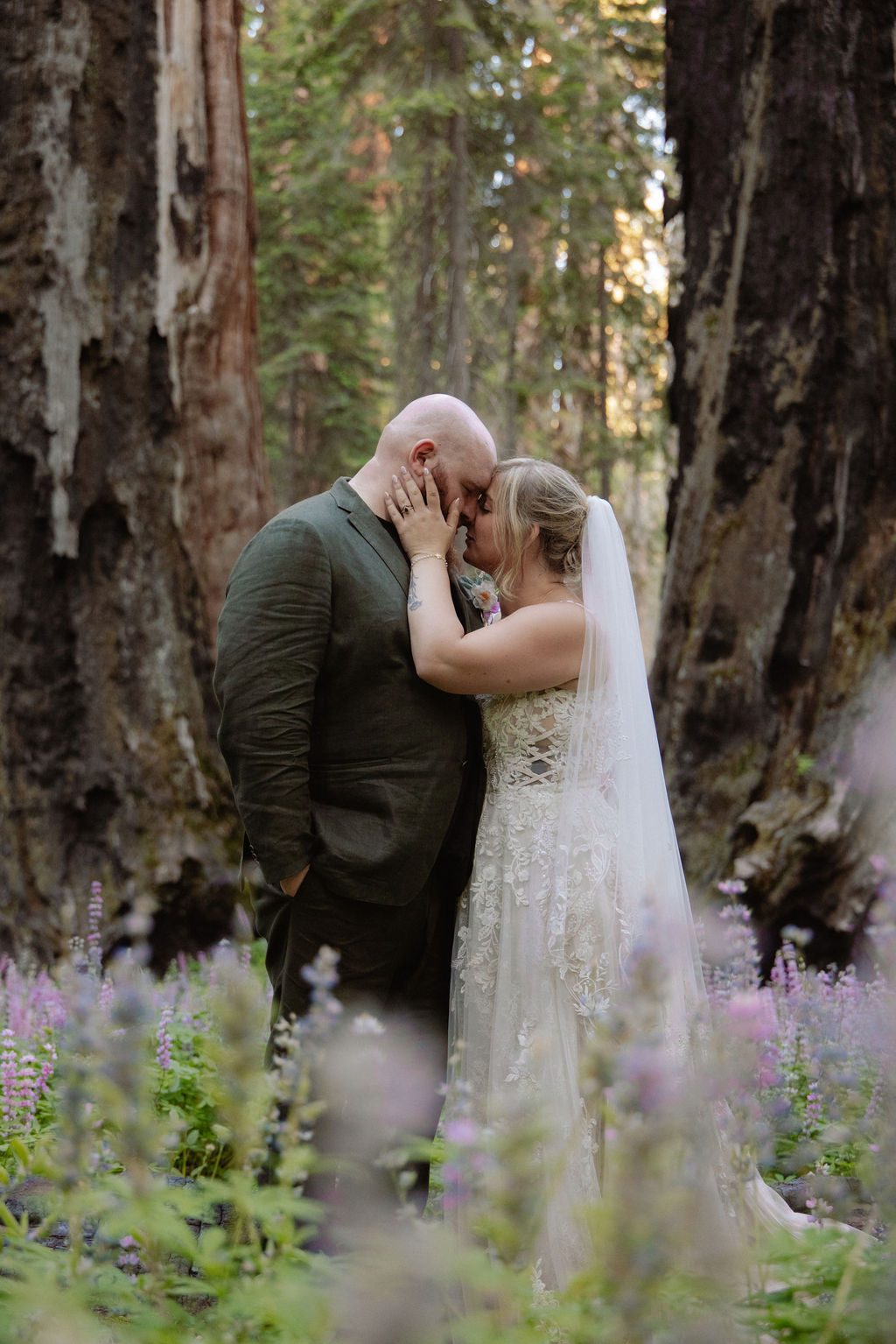 A bride and groom stand together in a forest clearing surrounded by tall trees and purple wildflowers.