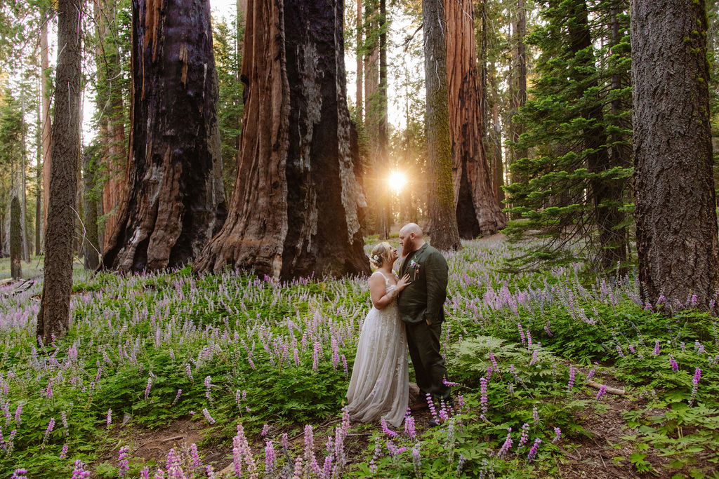 A bride and groom stand together in a forest clearing surrounded by tall trees and purple wildflowers.