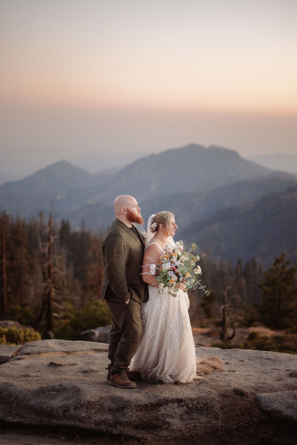 A couple in wedding attire stands on a rocky ledge, holding a bouquet, with mountains and trees in the background at sunset.