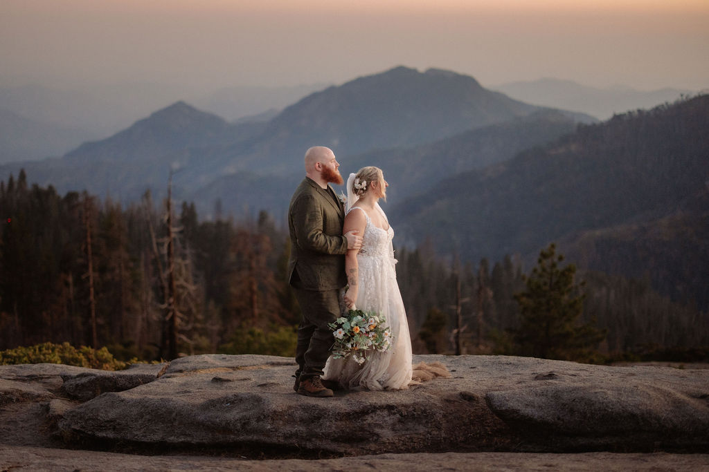 A couple in wedding attire stands on a rocky ledge, holding a bouquet, with mountains and trees in the background at sunset for a Yosemite and Sequoia elopement
