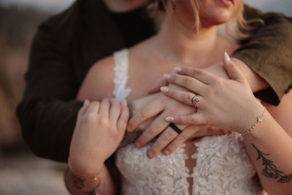 A couple in wedding attire stands on a rocky ledge, holding a bouquet, with mountains and trees in the background at sunset for a Yosemite and Sequoia elopement