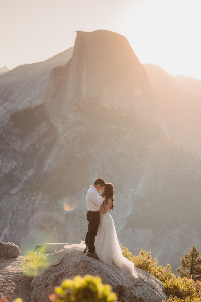 A couple in wedding attire stands on a rocky ledge, embracing, with a large mountain and soft sunlight in the background for your unique elopement