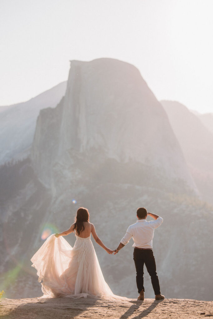 A couple in wedding attire stands on a rocky ledge, embracing, with a large mountain and soft sunlight in the background for your unique elopement
