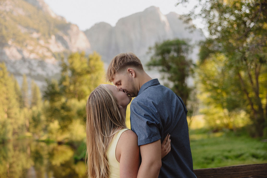 A couple stands outdoors, embracing and kissing, with a scenic mountain and tree landscape in the background.