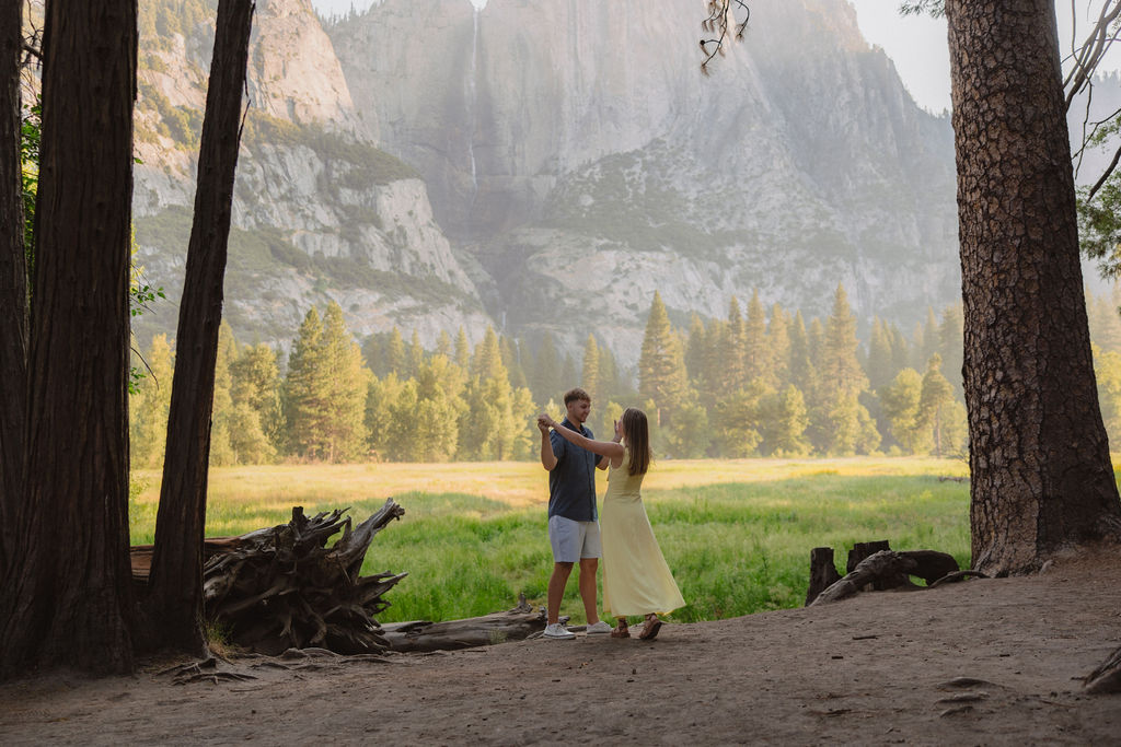 A couple stands outdoors, embracing and kissing, with a scenic mountain and tree landscape in the background.