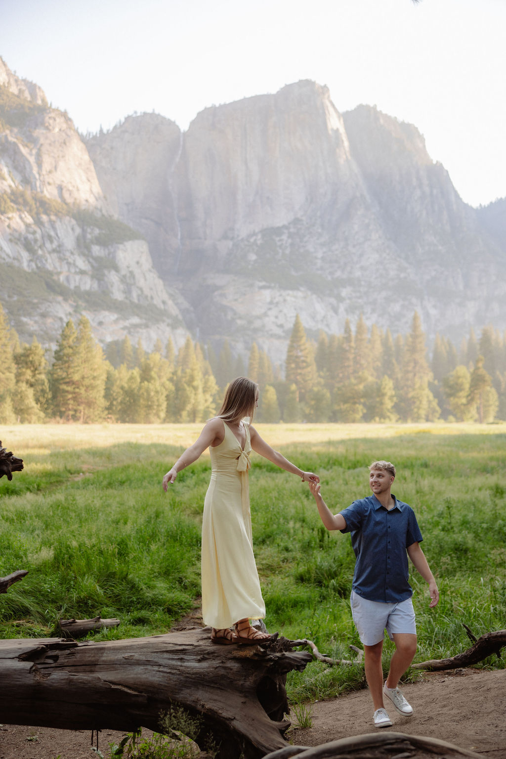 A woman in a yellow dress balances on a fallen tree while a man in shorts and a polo shirt holds her hand in a grassy valley with mountains in the background.