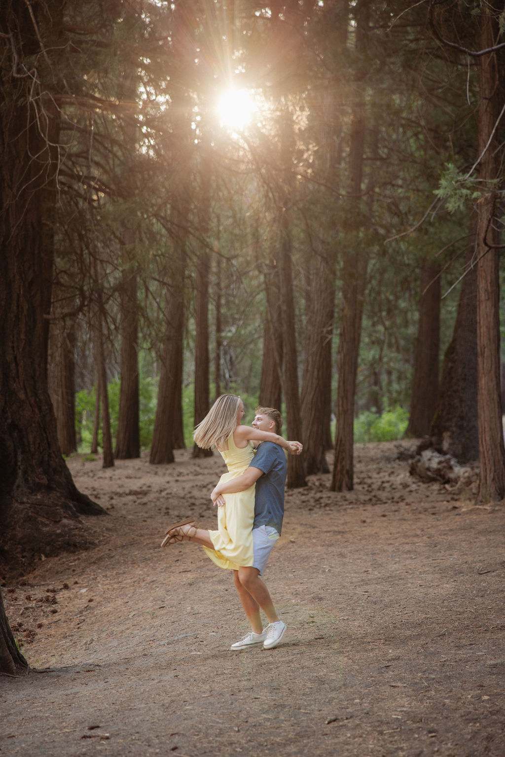 A woman in a yellow dress and a man in a blue shirt stand by a tree in a forest, looking at each other and smiling.