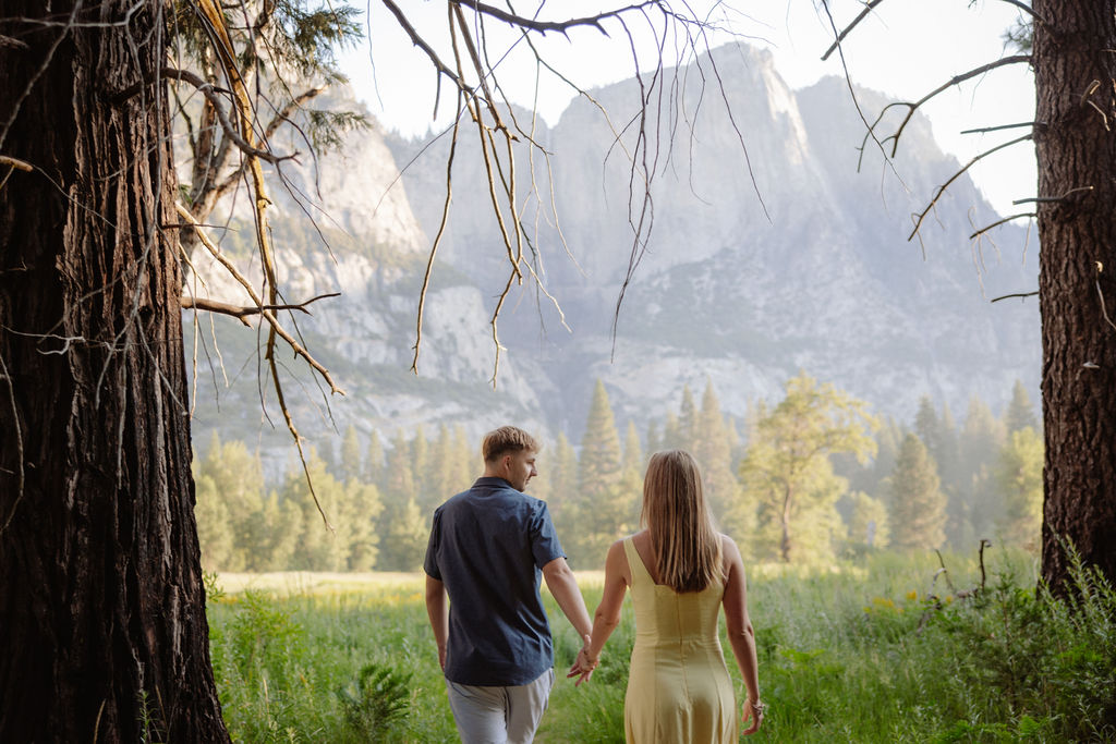 A couple stands outdoors, embracing with a scenic mountain and tree landscape in the background.