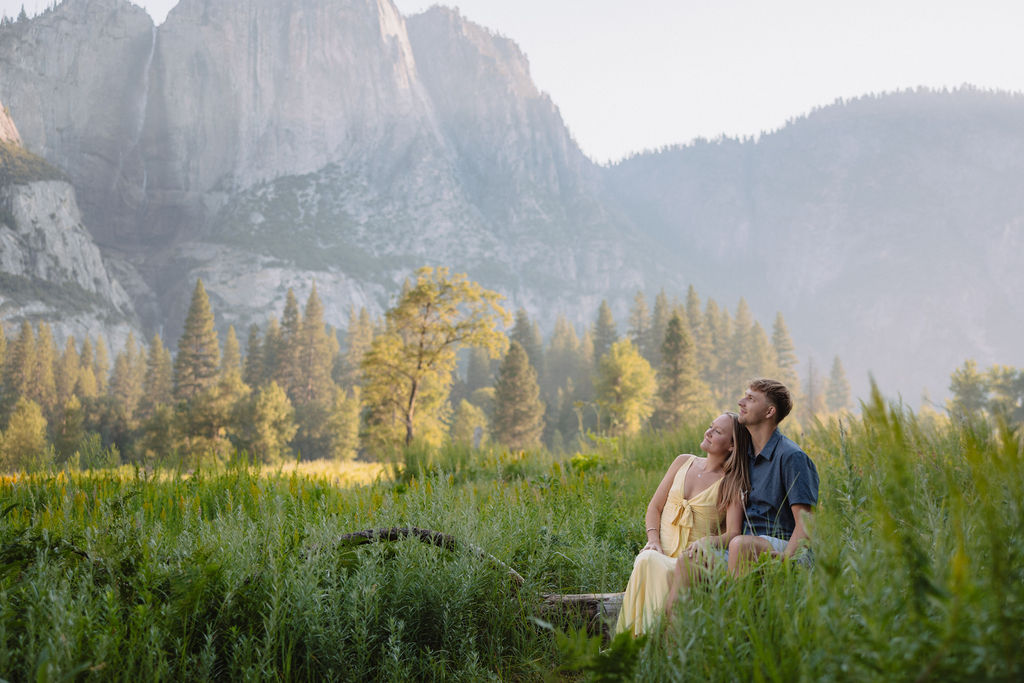 A couple stands outdoors, embracing with a scenic mountain and tree landscape in the background.