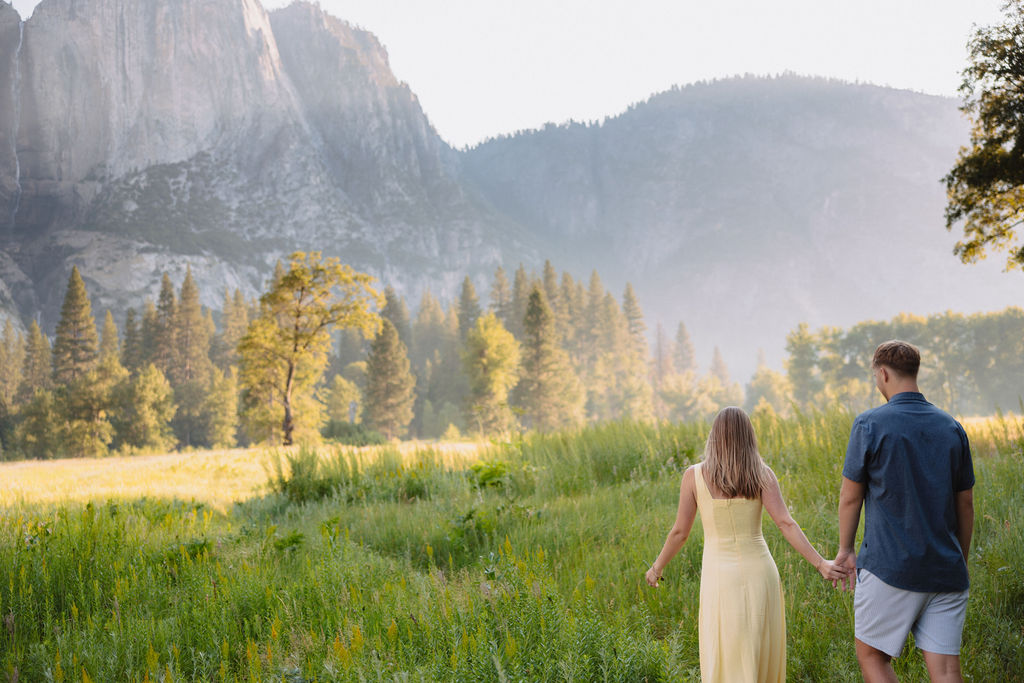 A couple stands outdoors, embracing with a scenic mountain and tree landscape in the background.