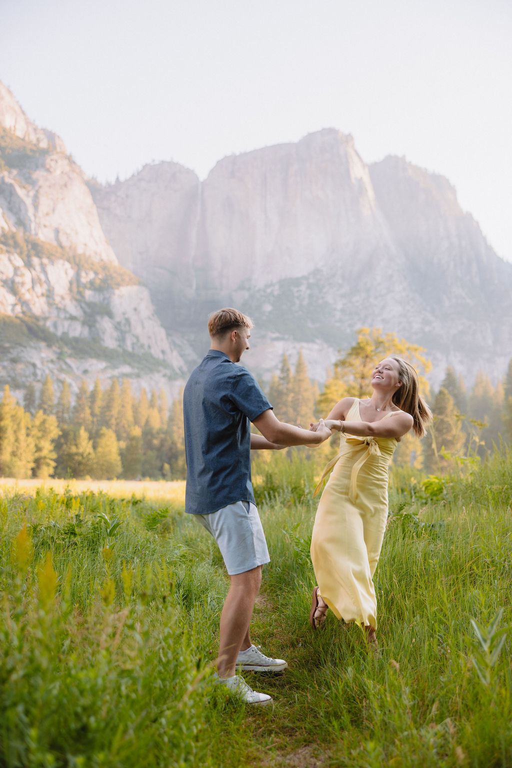 A woman in a yellow dress balances on a fallen tree while a man in shorts and a polo shirt holds her hand in a grassy valley with mountains in the background.
