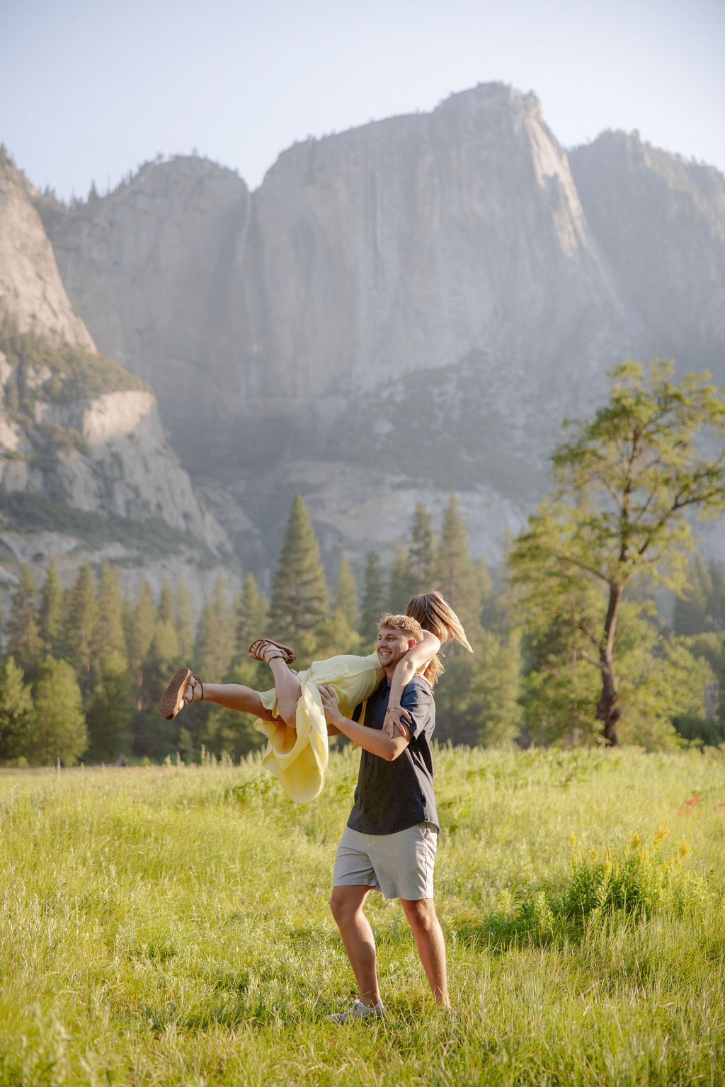 A couple stands outdoors, embracing with a scenic mountain and tree landscape in the background.