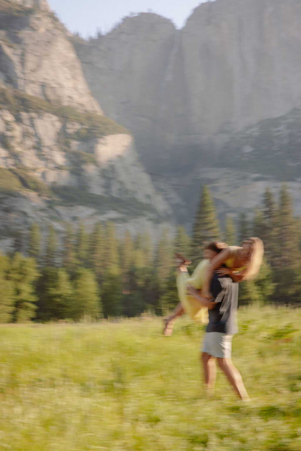 A couple stands outdoors, embracing with a scenic mountain and tree landscape in the background.