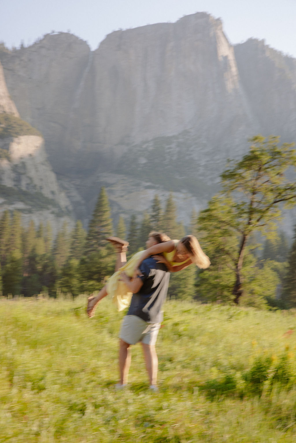 A woman in a yellow dress balances on a fallen tree while a man in shorts and a polo shirt holds her hand in a grassy valley with mountains in the background.